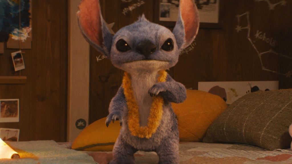 Live-action Stitch from Disney's Lilo and Stitch (2025) remake, wearing a yellow lei and looking mischievous on a bed.