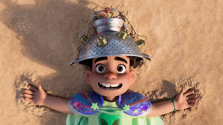The main character from Pixar's Elio lies on the sand, looking up at the camera while wearing a homemade helmet made from a colander.