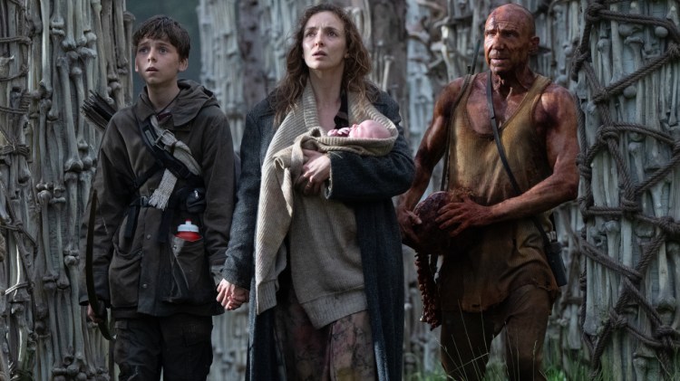 Characters in 28 Years Later walk through a bone structure while holding a baby.