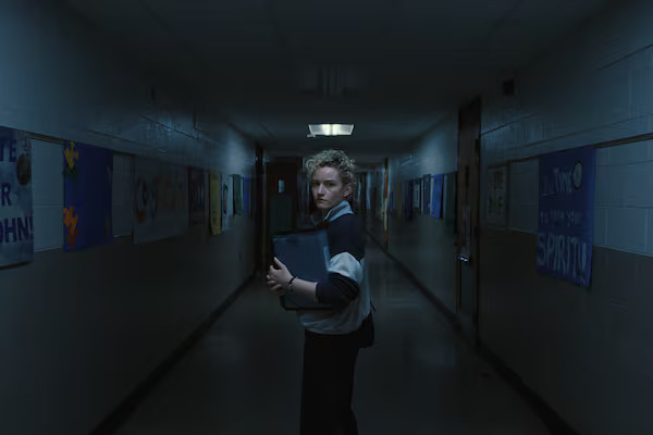 Julia Garner looks back with a worried expression while standing alone in a dark school hallway in the horror film Weapons.